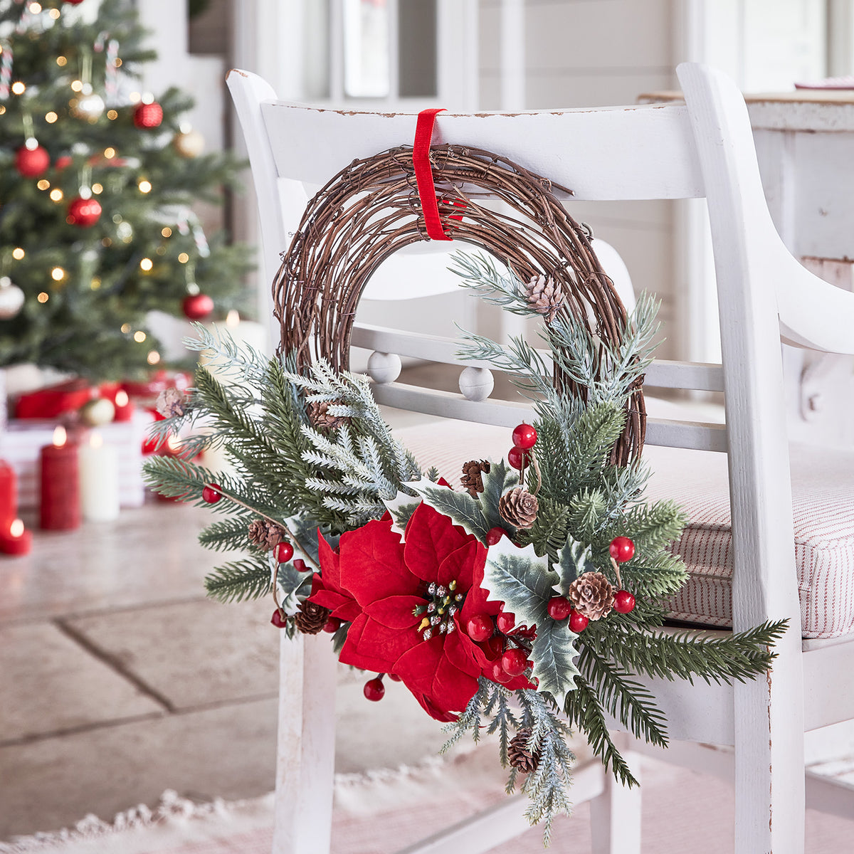Weihnachtskranz mit roten Sternblüten und Tannenzweigen hängt an weißem Stuhl vor Weihnachtsbaum