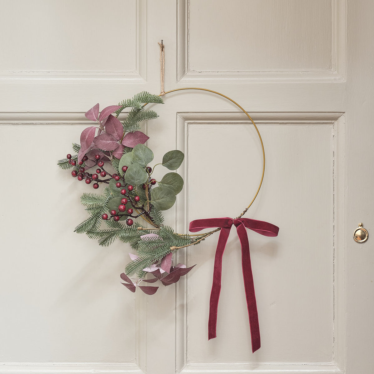 Minimalistischer Weihnachtskranz mit rotem Samtband und Kunstlaub, hängend an weißer Tür