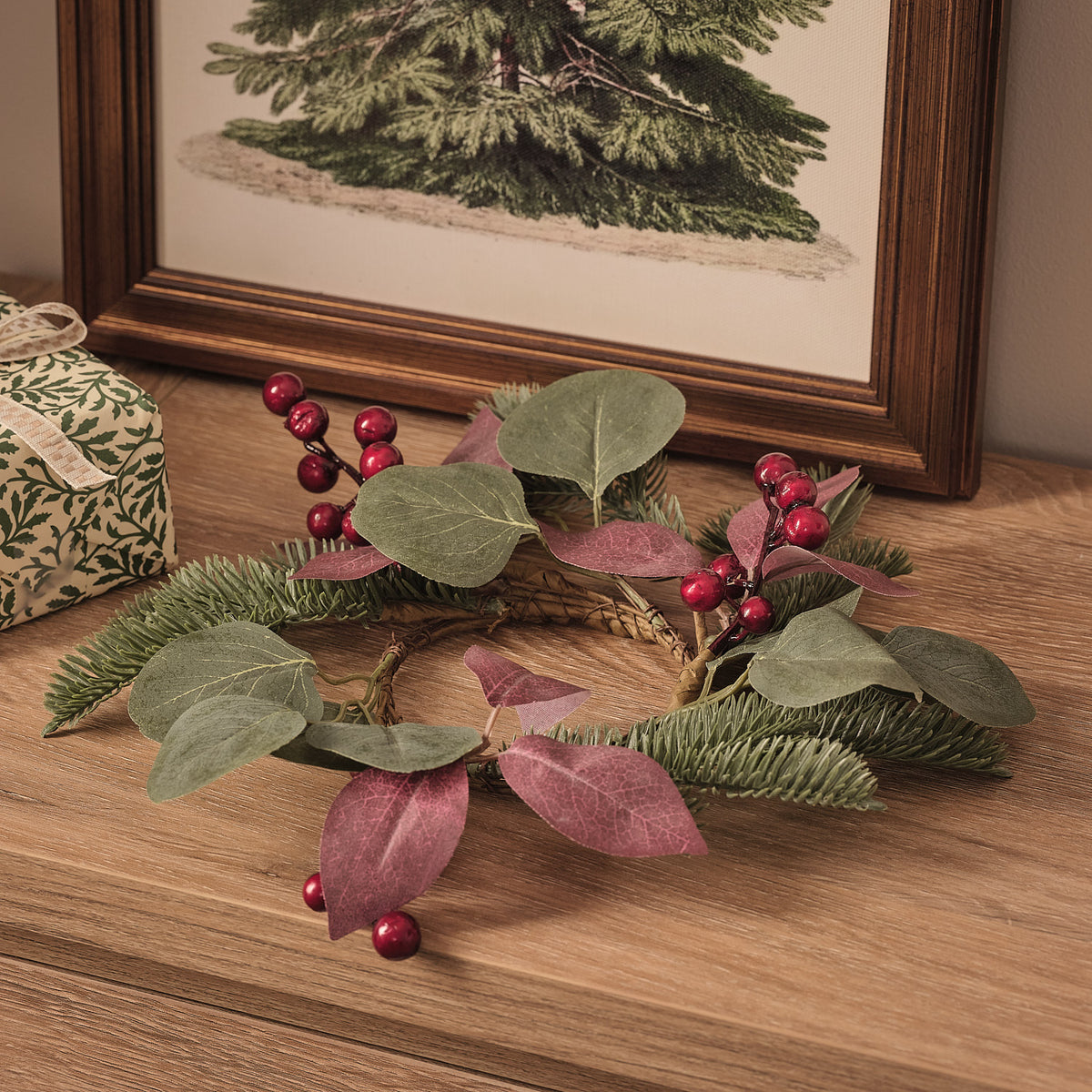 Weihnachtlicher Kerzenring mit rotem Beeren- und grünem Kunstlaub auf Holzfläche vor Bilderrahmen