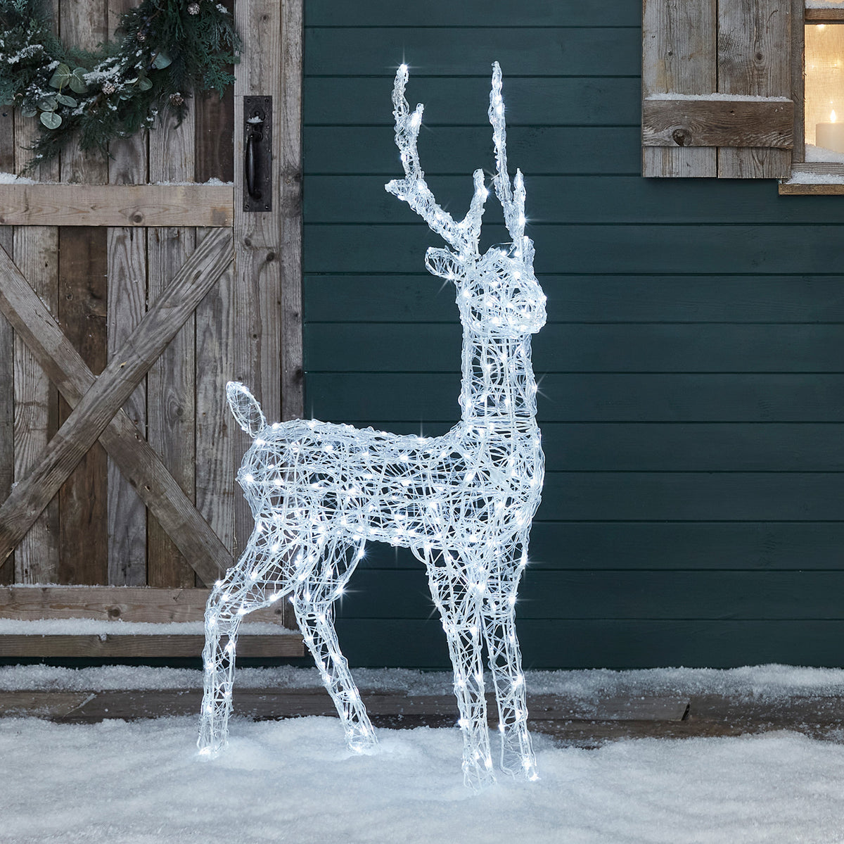 Leuchtendes Acryl-Rentier mit weißen LEDs steht auf Schnee vor einer Holztür