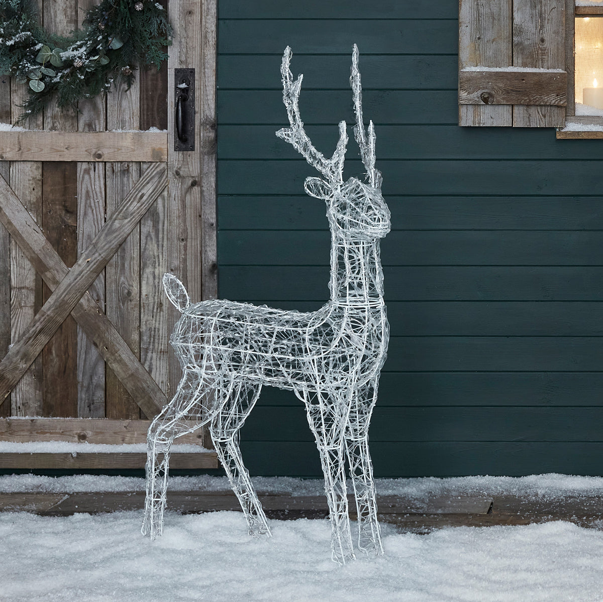 Acryl-Rentier mit weißen LEDs vor Holztür und Fenster mit Kerzenlicht im Schnee