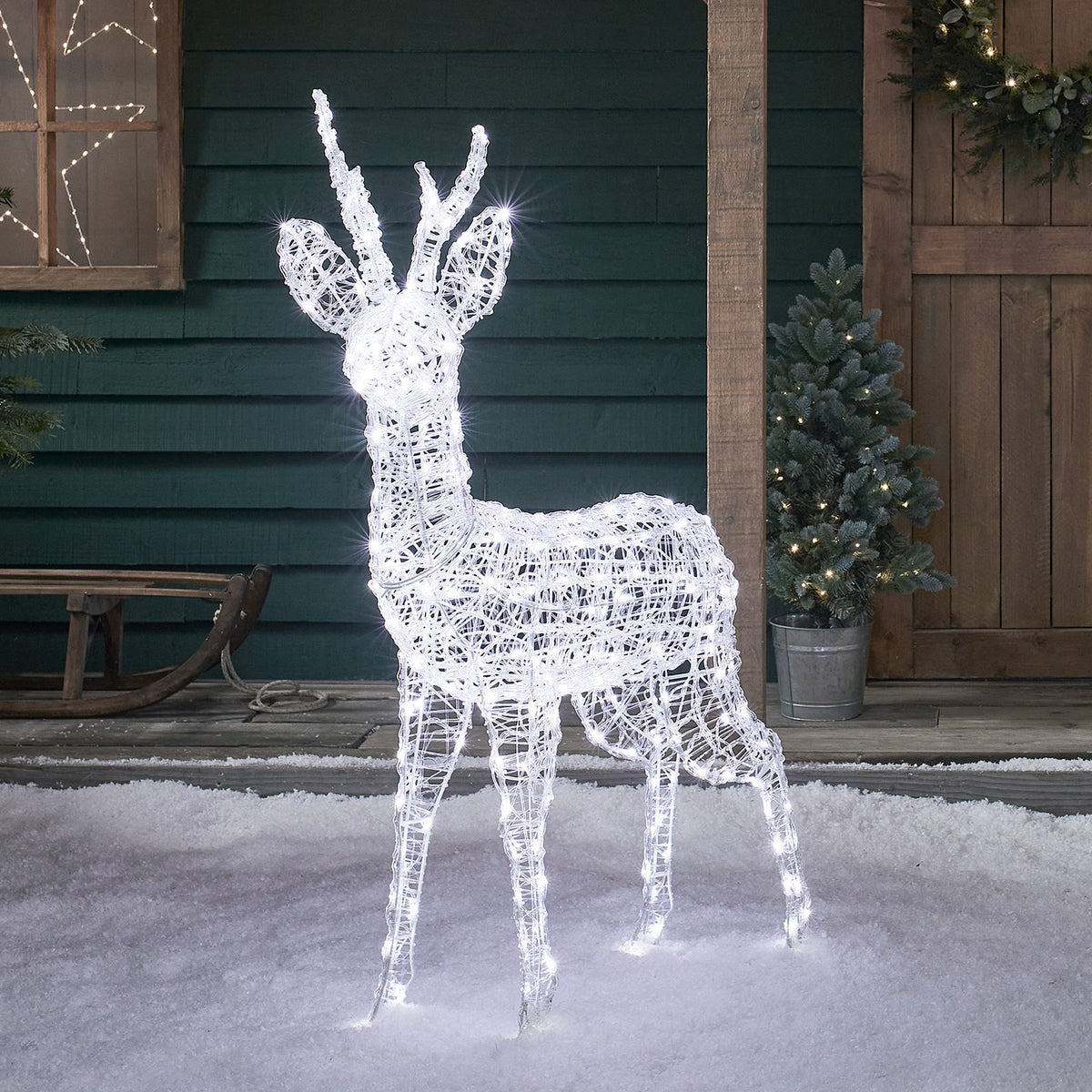 Acryl-Rentier mit kalten weißen LEDs steht auf Schnee neben Holztür und Tannenzweig