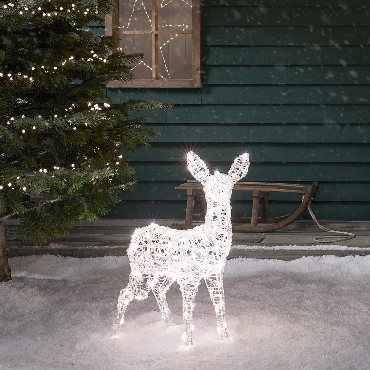 LED-Rehkitz aus Acryl mit weißem Licht vor grünem Holzhaus und Schlitten im Schnee