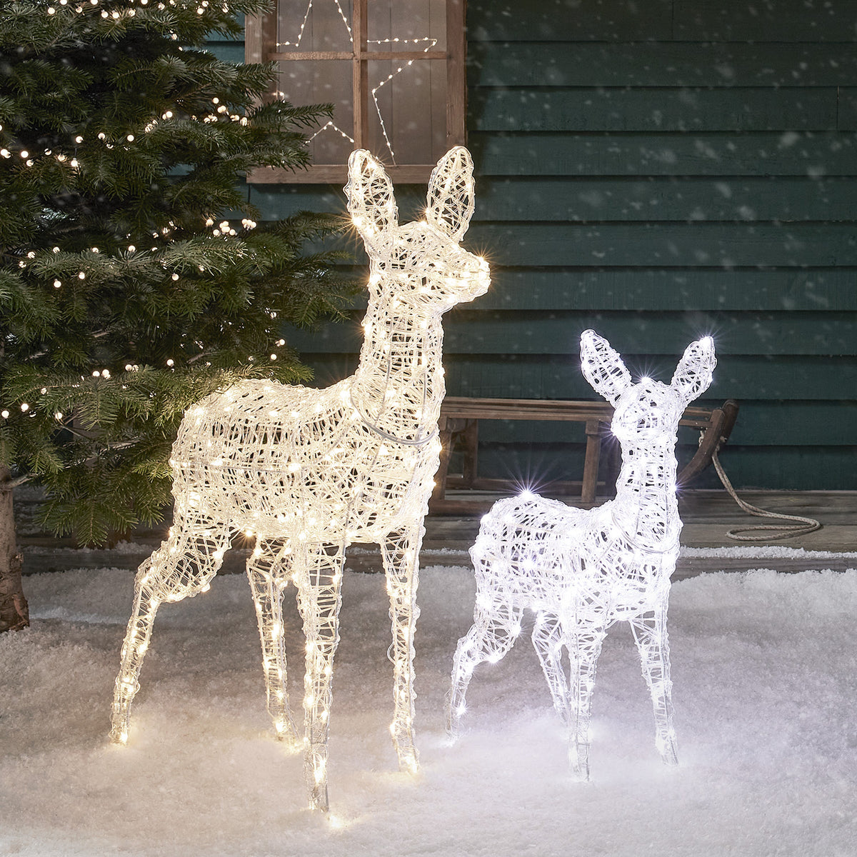 LED-Rentiere mit warmweißen und weißen Lichtern auf Schnee vor dunklem Holzhaus mit Fenster