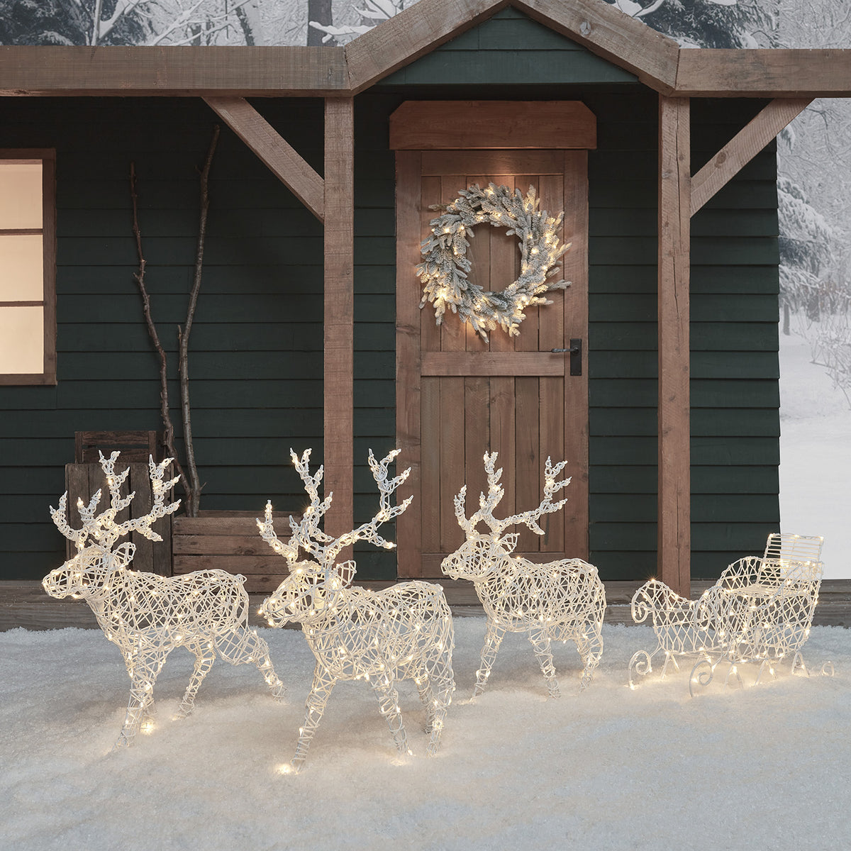 Drei beleuchtete LED-Rentiere mit Schlitten vor einer Holztür im Schnee