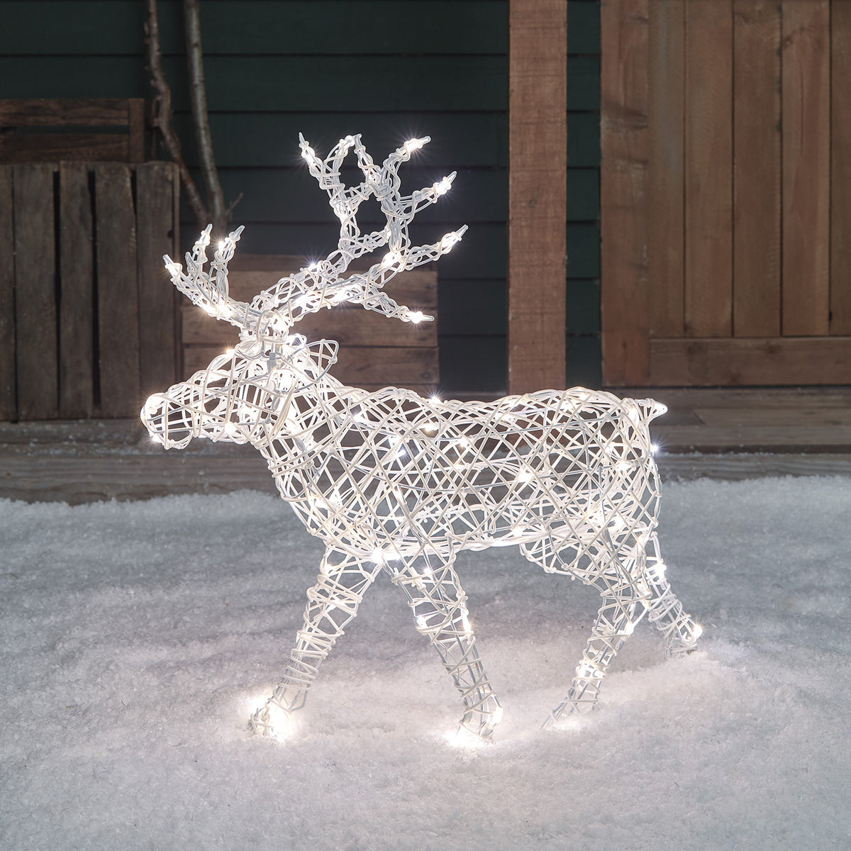 LED-Rentier mit weißem Kunststoffgeflecht und weißen LEDs stehend im Schnee vor Holzwand