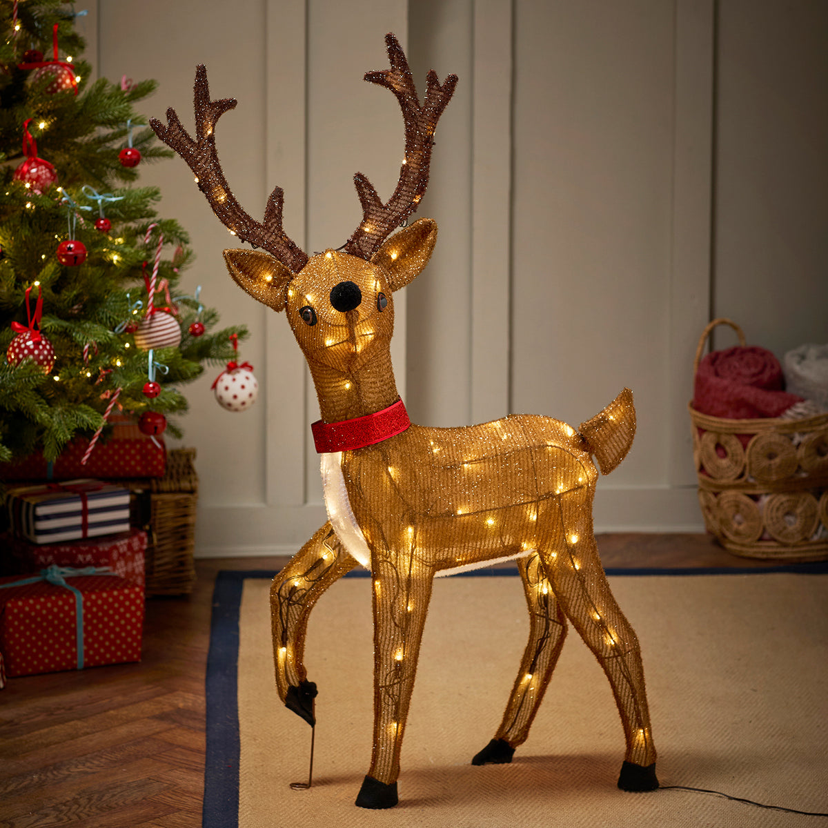 LED-Rentier mit rotem Schal neben geschmücktem Weihnachtsbaum und Geschenken im Wohnzimmer