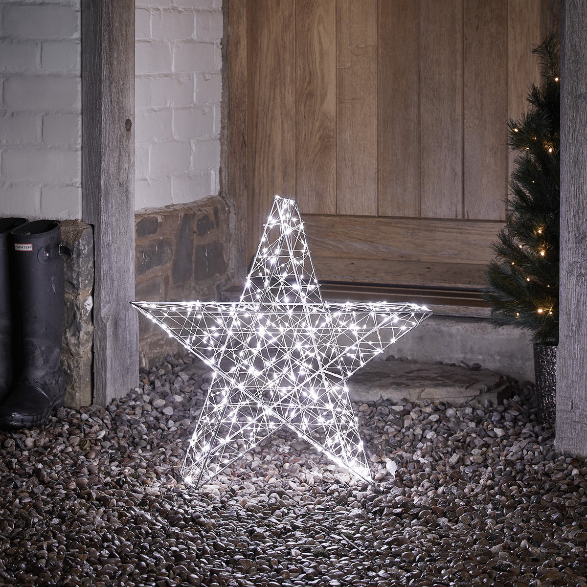 3D Stern mit kaltweißen LEDs beleuchtet auf Kies vor Holztür und Tannenzweig