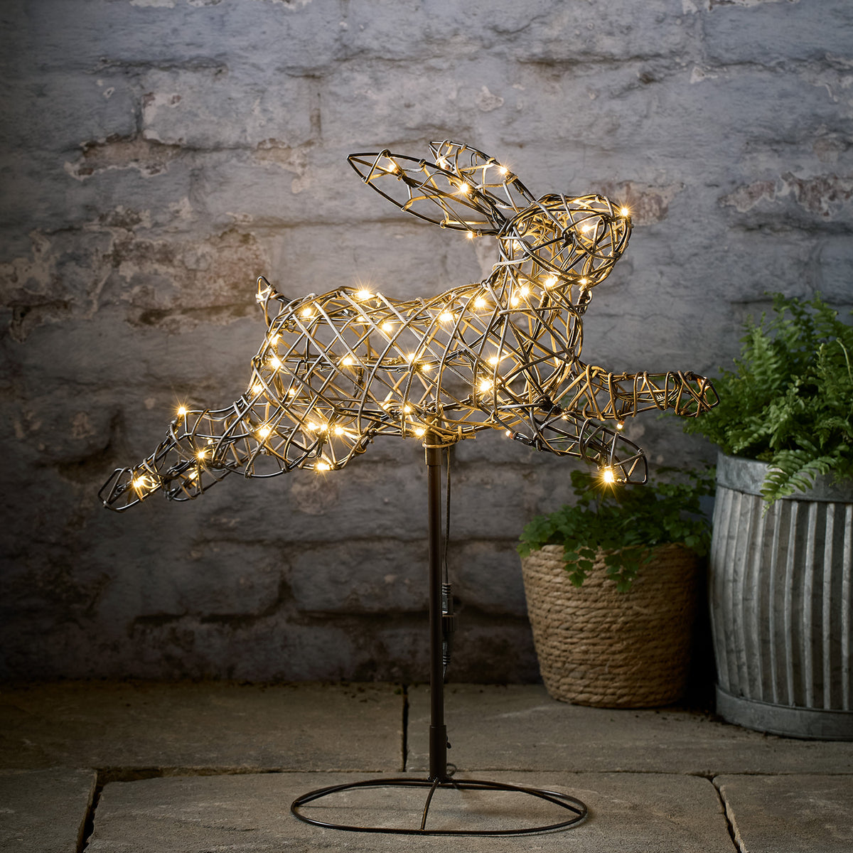 Leuchtender LED-Hase aus Rattanimitat mit 60 warmweißen LEDs auf Metallständer vor Steinwand