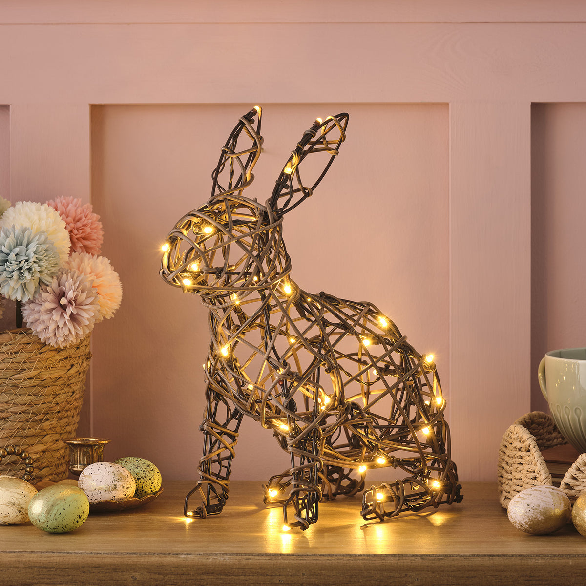 Beleuchteter Rattan-Hase auf Tisch mit Osterdeko und rosa Wand im Hintergrund