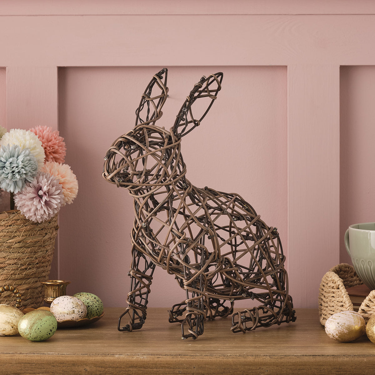 Unbeleuchteter Rattan-Hase auf Tisch mit Osterdeko vor rosa Wand