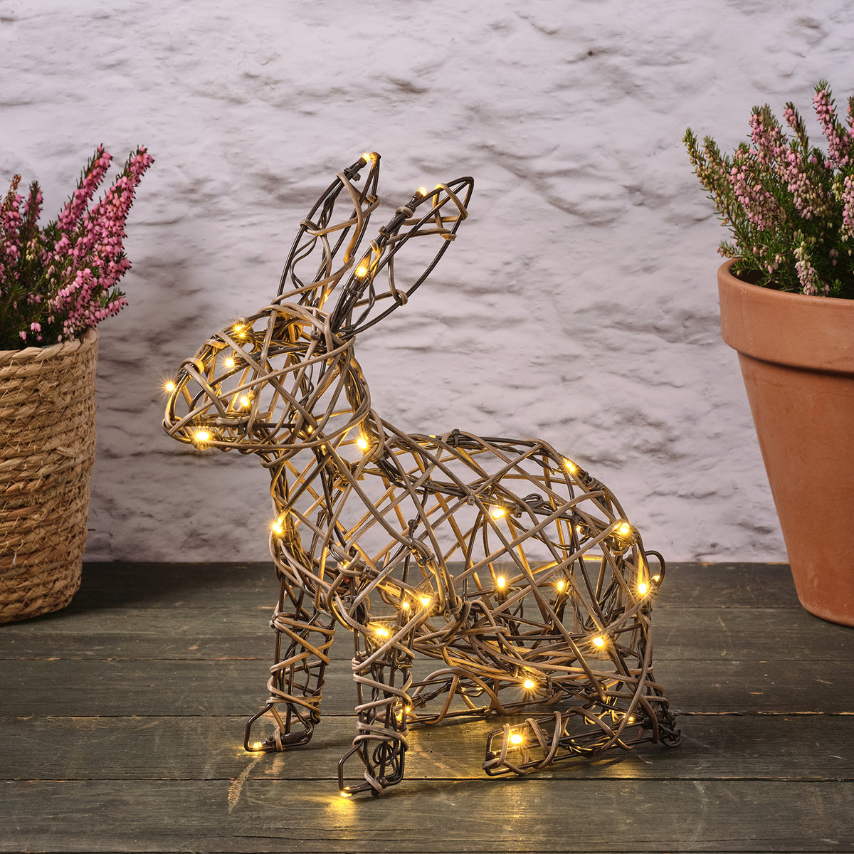 Rattan-Hase mit warmweißen LEDs auf dunklem Holzboden zwischen zwei Blumentöpfen