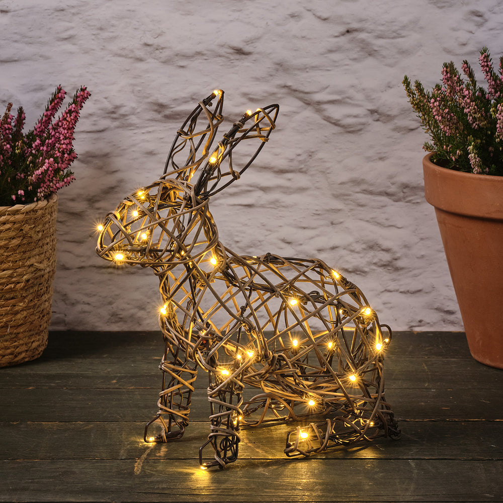 Rattan-Hase mit warmweißen LEDs auf dunklem Holzboden zwischen zwei Blumentöpfen