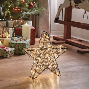 Beleuchteter Rattan-LED-Stern vor Weihnachtsbaum und Geschenken auf Holzboden