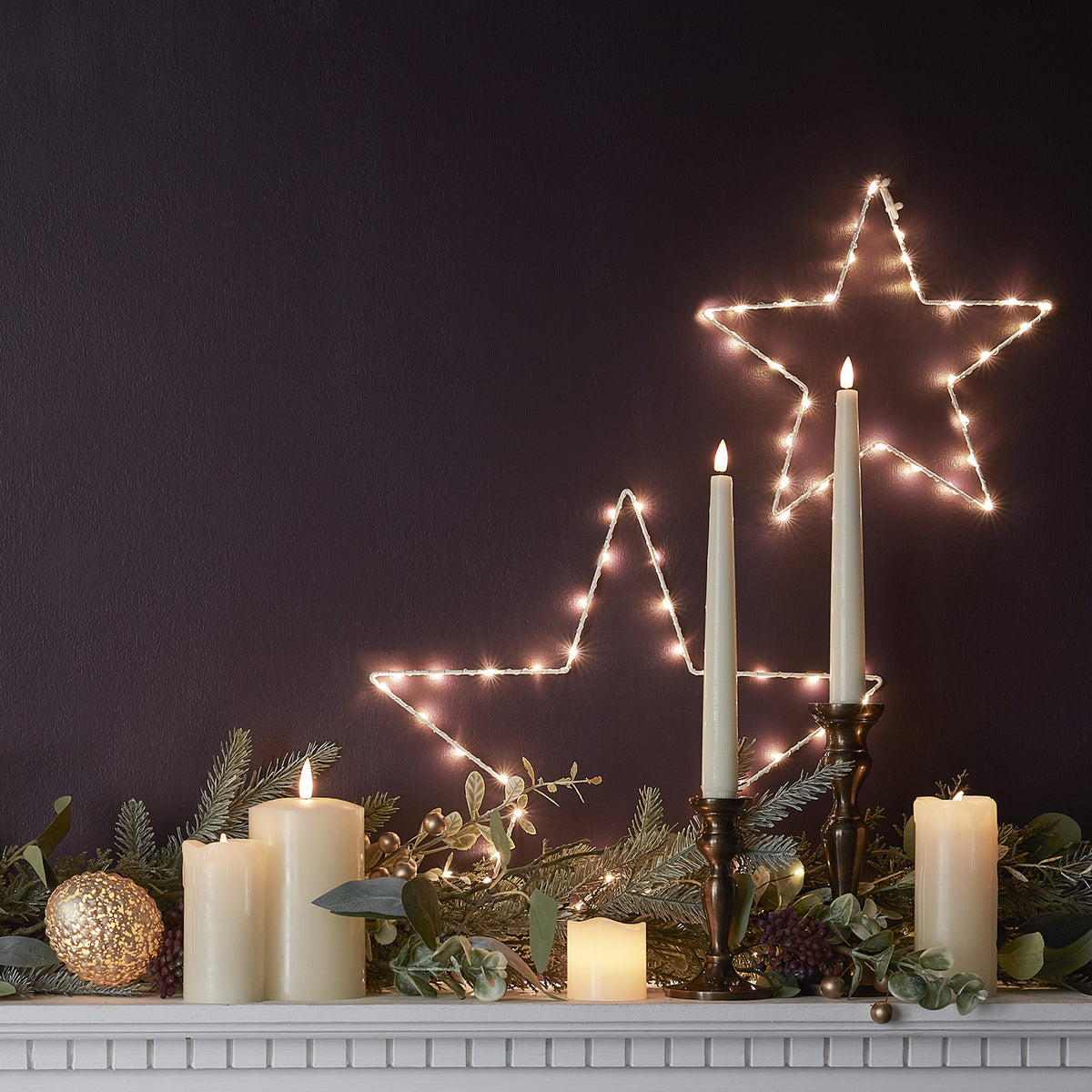 Beleuchtete LED-Drahtsterne an dunkler Wand mit brennenden Kerzen und Tannenzweigen