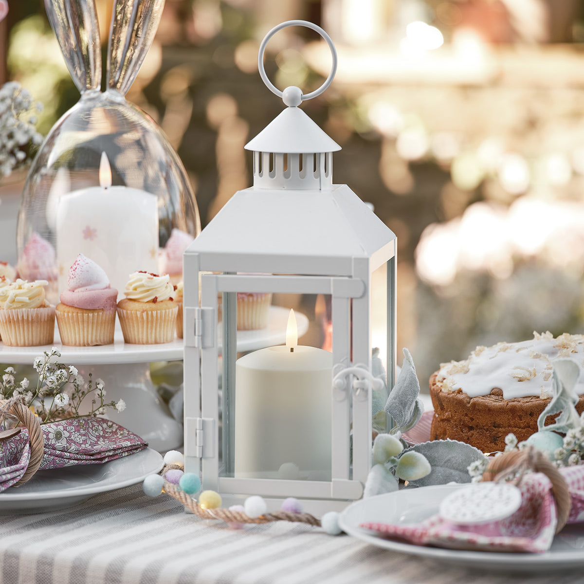Weiße Laterne mit LED-Kerze auf gedecktem Tisch mit Kuchen und Cupcakes