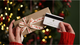 Gift box with a decorative bow and a gift card from one Stop German Shop held by hands against a festive background.