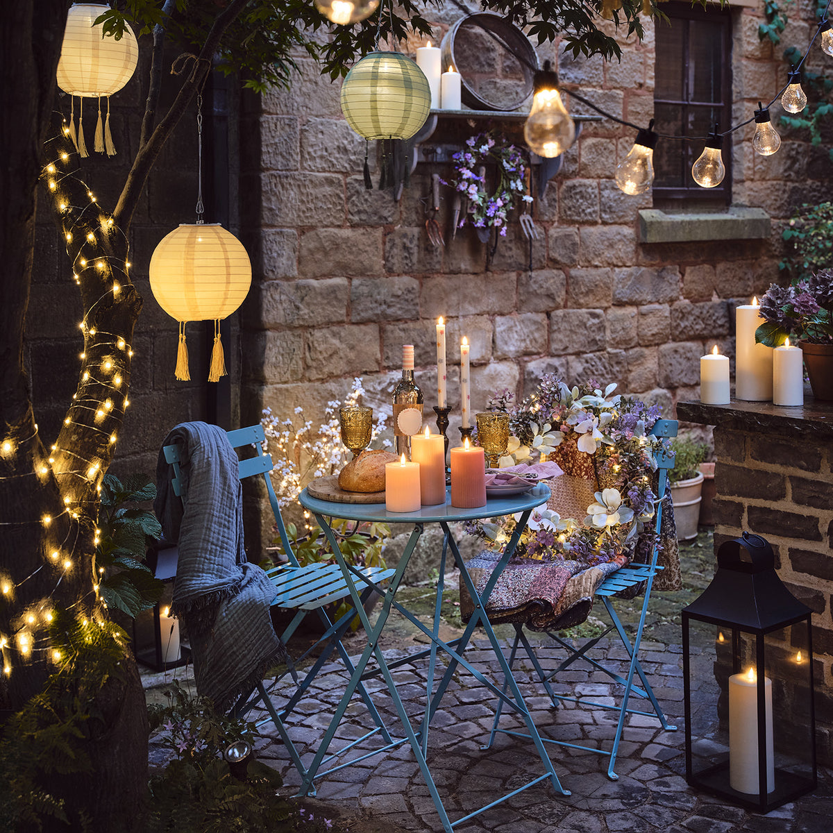 Beleuchtete Solar-Lampion hängen über einem kleinen Gartentisch mit Kerzen und Blumen