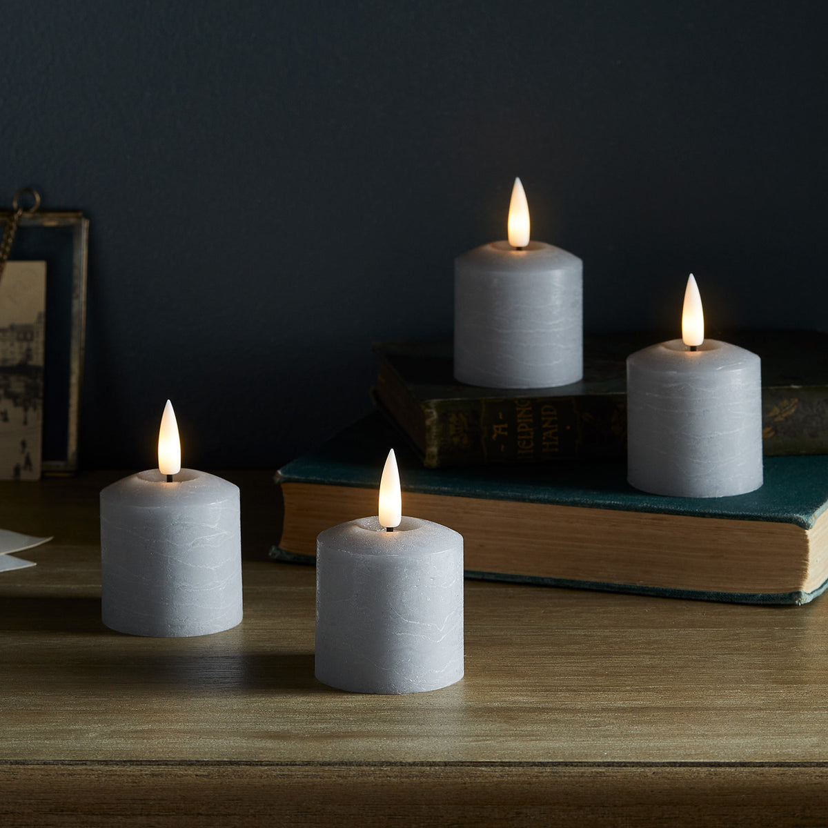 Vier graue LED Votivkerzen mit leuchtenden Flammen auf Holztisch vor dunkler Wand und Büchern