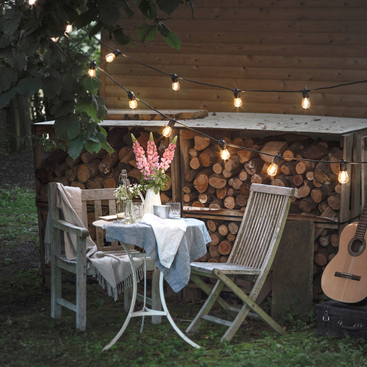 Garten mit Lichterkette, Holztisch, Stühlen und Blumen im Vordergrund
