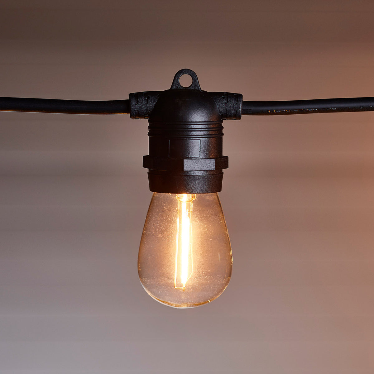 Nahaufnahme einer warmweiß leuchtenden Glühbirne an schwarzem Kabel vor weißer Decke