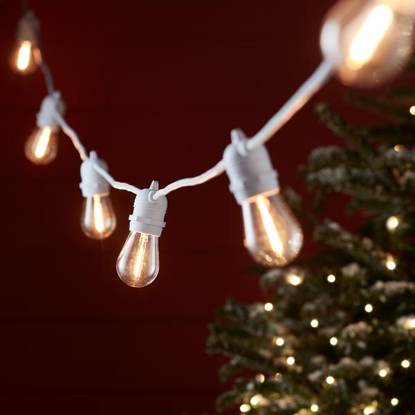 Warmweiße Glühbirnen an weißem Kabel vor rotem Hintergrund und Weihnachtsbaum