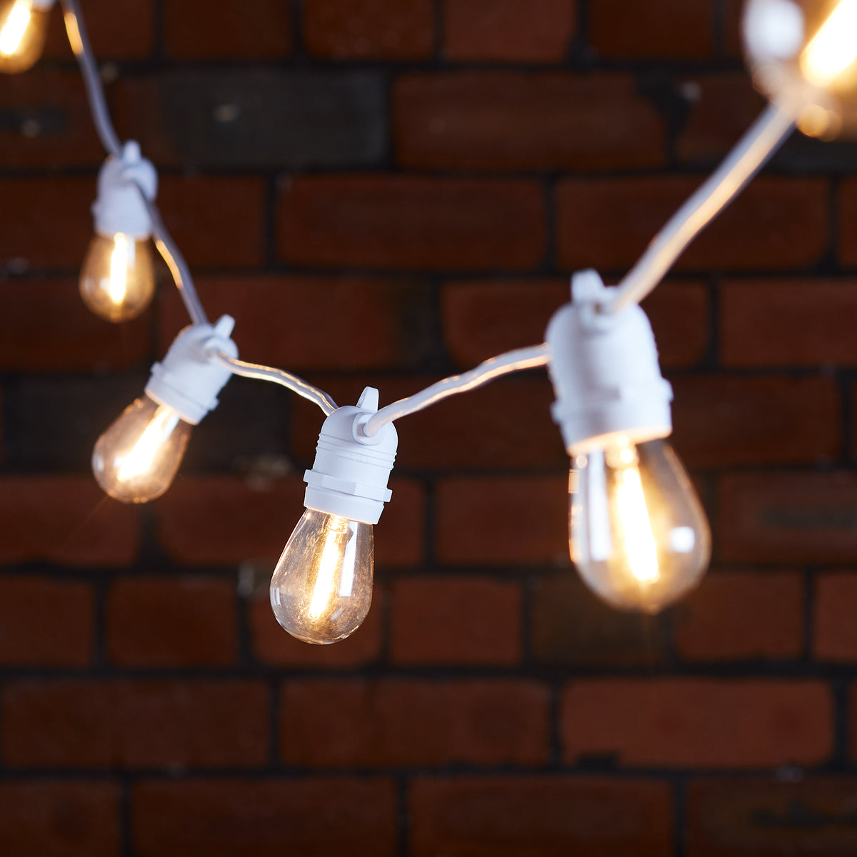 Warmweiße Glühbirnen an weißem Kabel vor roter Backsteinwand, Lichterkette im Fokus