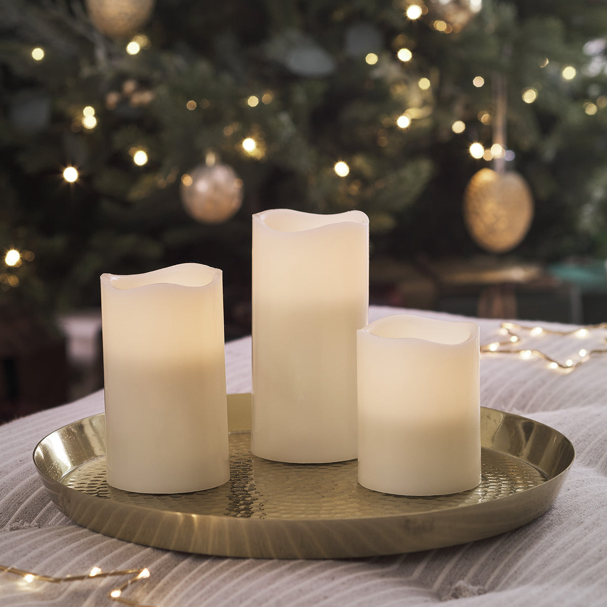 Drei Echtwachs LED Kerzen auf goldenem Tablett vor einem Weihnachtsbaum mit Lichterkette