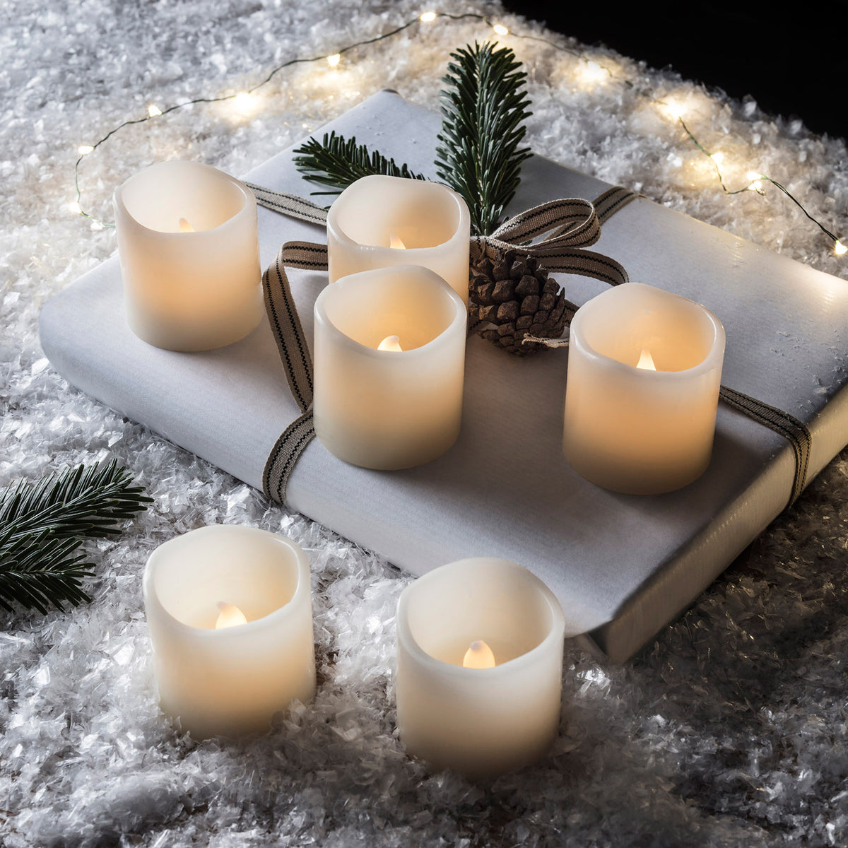Sechs LED Votivkerzen auf Geschenk mit Tannenzweig und Zapfen, umgeben von Kunstschnee und Lichterkette