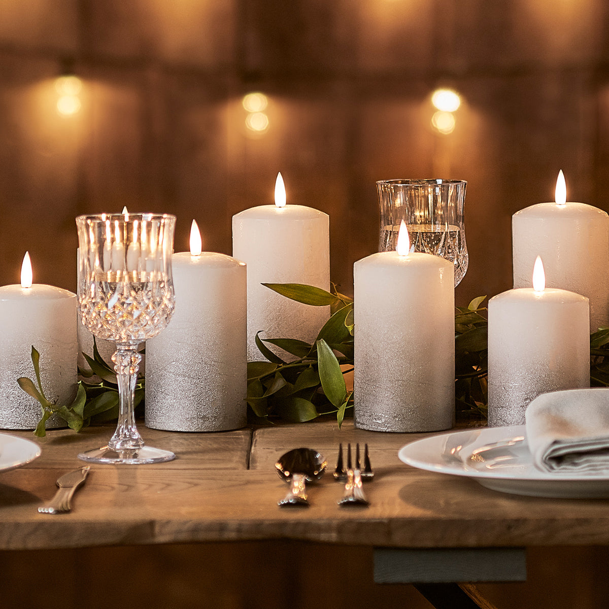 Mehrere graue Ombré LED-Kerzen und Kristallgläser auf gedecktem Holztisch mit Lichterkette