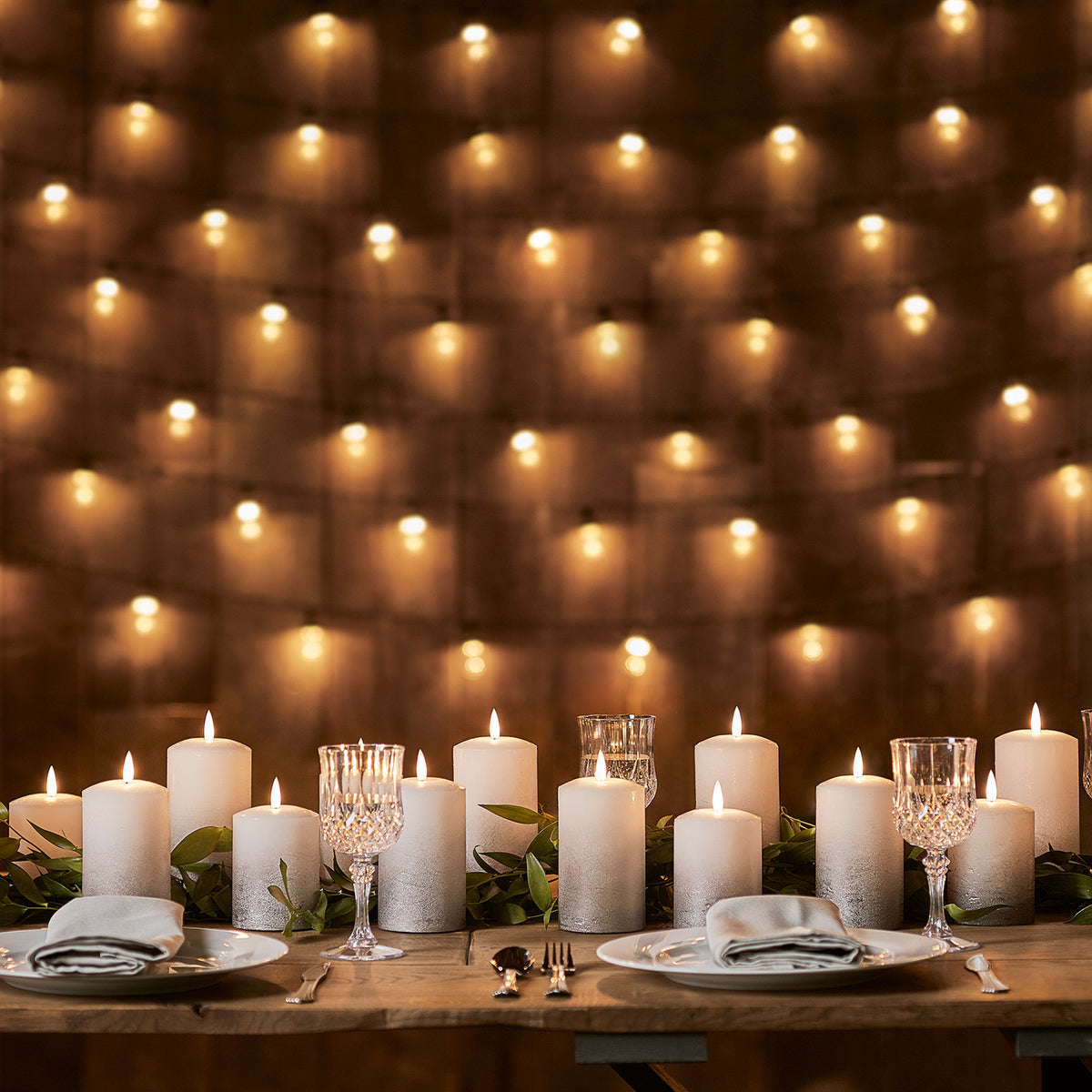 Viele graue Ombré LED-Kerzen und Kristallgläser auf Holztisch vor Wand mit warmen Lichtern