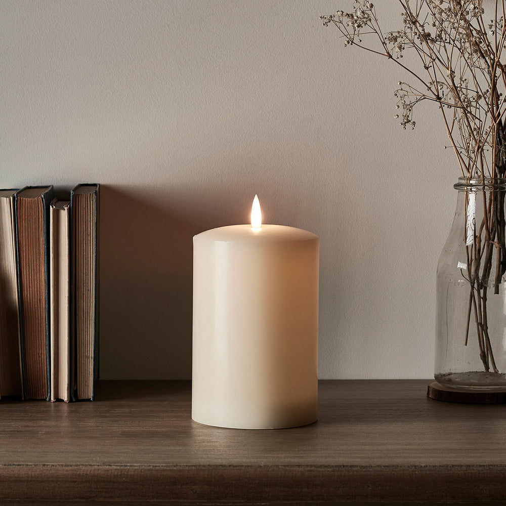 Breite weiße LED Stumpenkerze mit flackernder Flamme auf Holzregal neben Büchern und Vase