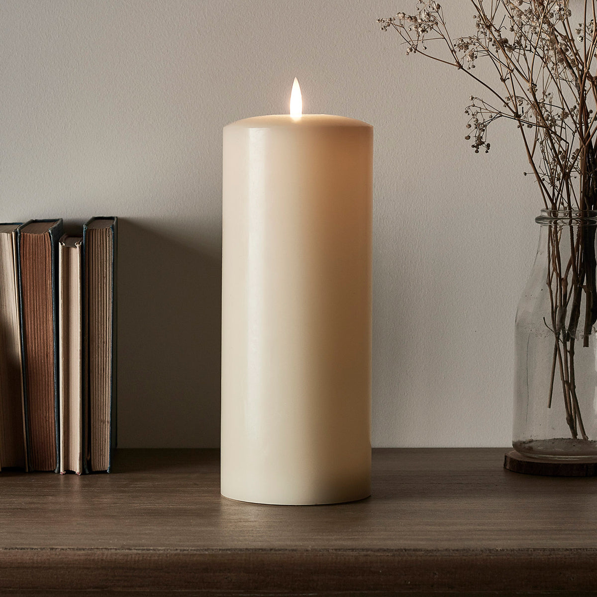 Breite TruGlow LED Stumpenkerze mit flackernder LED-Flamme auf Holzfläche neben Büchern und Vase