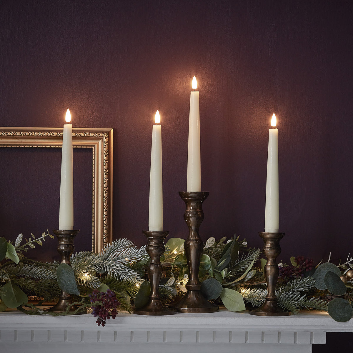 Vier TruGlow LED Stabkerzen in antiken Kerzenhaltern vor dunkler Wand mit Weihnachtsdeko
