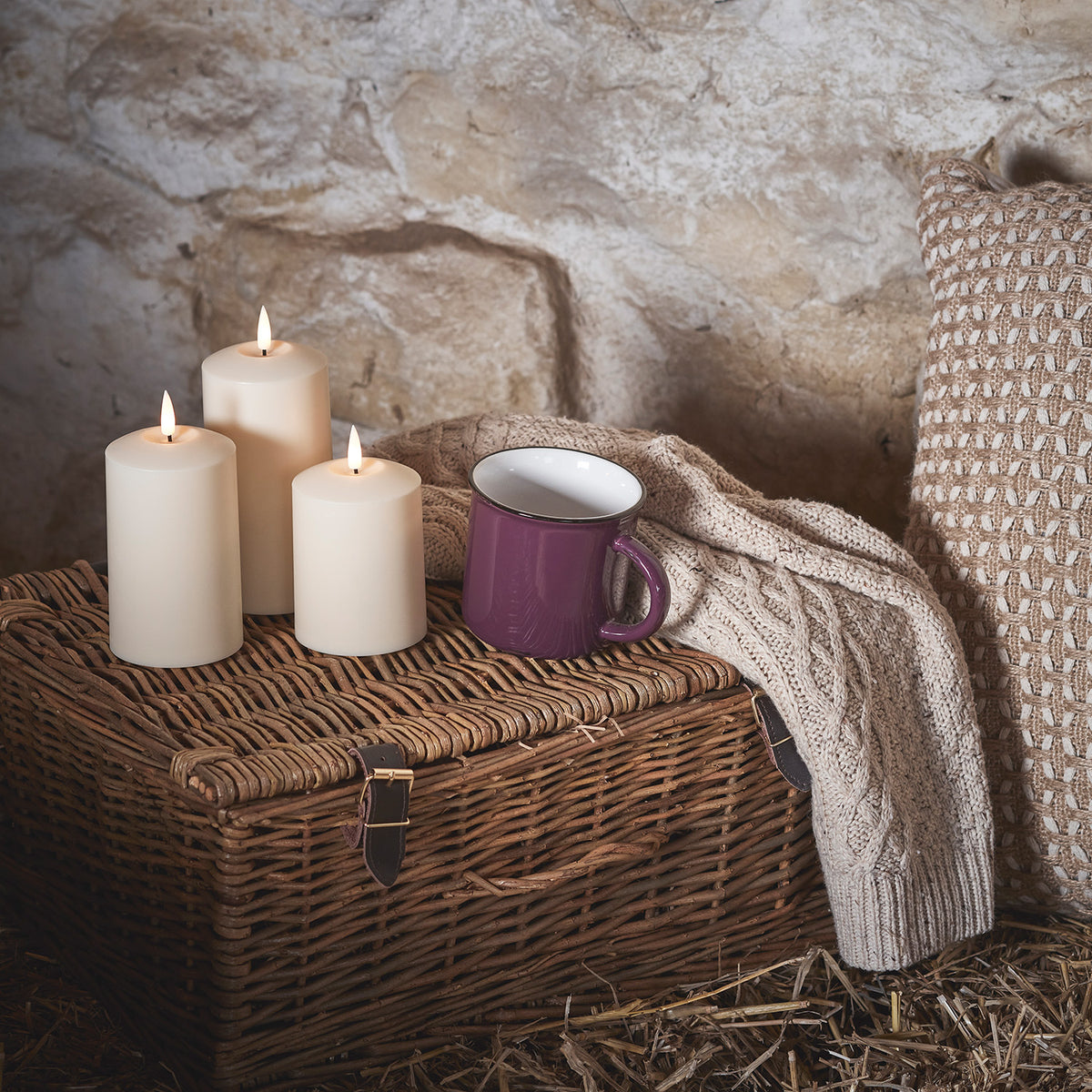 Drei brennende TruGlow LED Kerzen auf geflochtenem Korb neben lila Tasse und Strickdecke