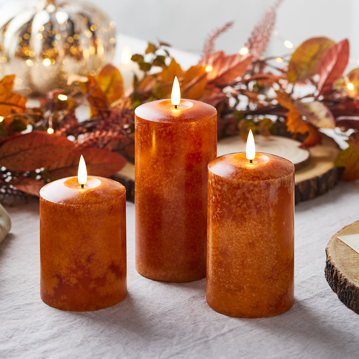 Trio orangefarbener LED Echtwachskerzen mit herbstlicher Deko im Hintergrund auf hellem Stoff