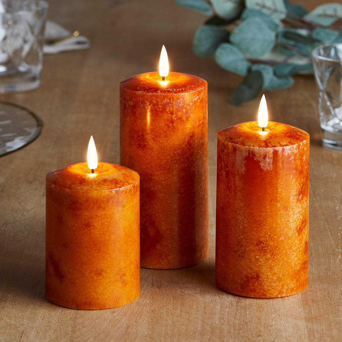 Drei orangefarbene LED Echtwachskerzen auf hellem Holztisch mit Gläsern und grünen Blättern
