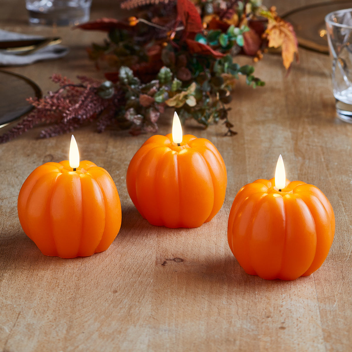 Drei orangefarbene LED-Kerzen in Kürbisform auf einem Holztisch mit herbstlicher Dekoration im Hintergrund