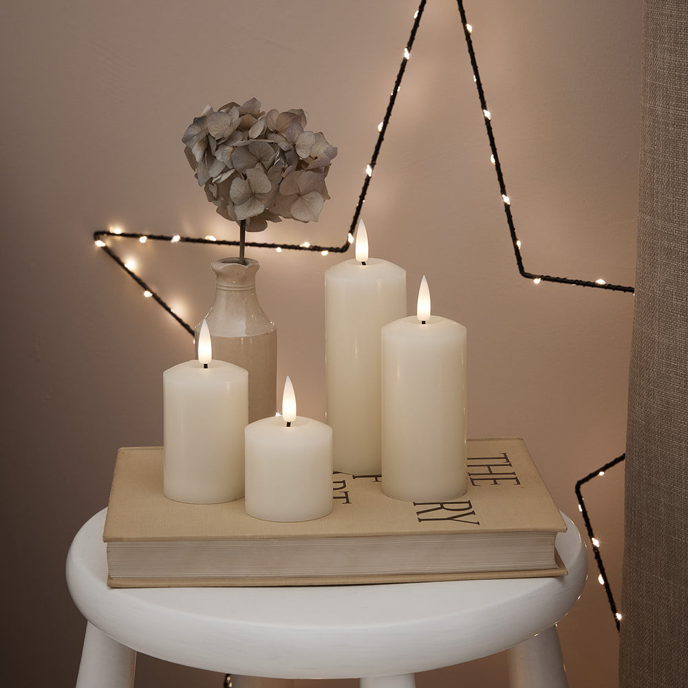 Vier elfenbeinfarbene LED Stumpenkerzen auf einem Buch mit getrockneter Blume und Lichterkette im Hintergrund