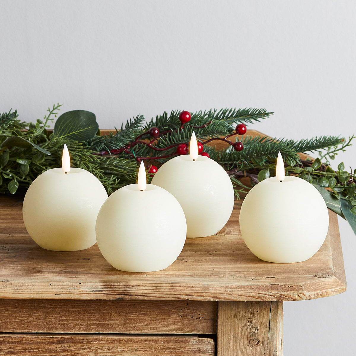 Vier runde LED-Kerzen aus echtem Wachs mit Weihnachtsgrün und roten Beeren auf Holzbank