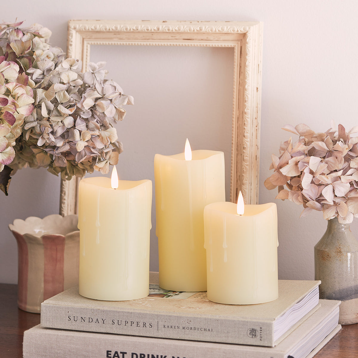 Drei brennende LED Stumpenkerzen auf Büchern mit getrockneten Blumen und Bilderrahmen