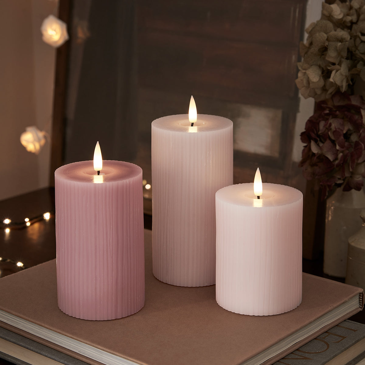 Drei brennende LED-Kerzen in Rosa- und Pastelltönen auf Buch vor Spiegel und getrockneten Blumen