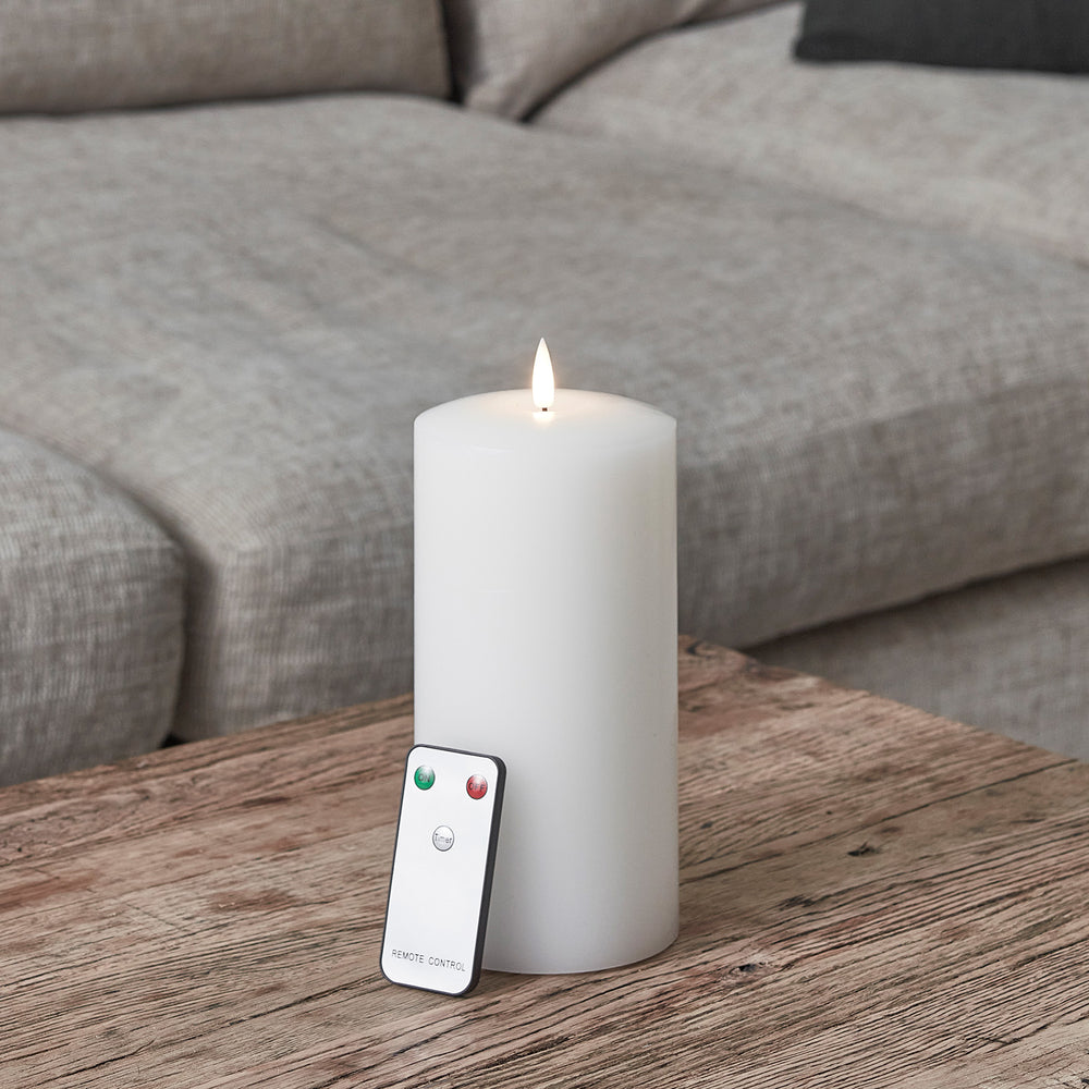 Weiße LED Stumpenkerze mit Flamme und Fernbedienung auf Holztisch vor grauem Sofa