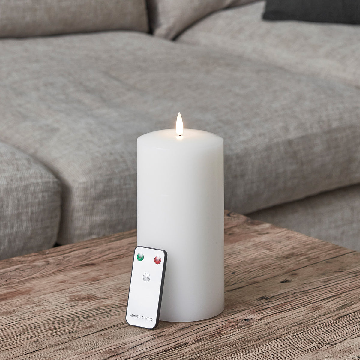 Weiße LED Stumpenkerze mit Flamme und Fernbedienung auf Holztisch vor grauem Sofa