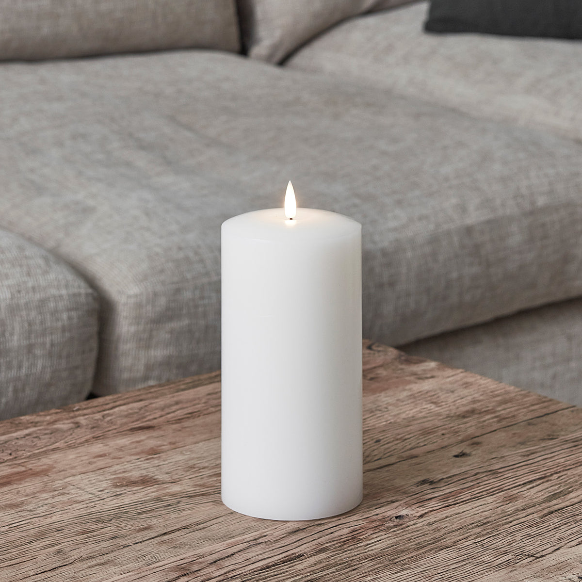 Weiße breite LED Stumpenkerze aus Echtwachs auf Holztisch vor grauem Sofa