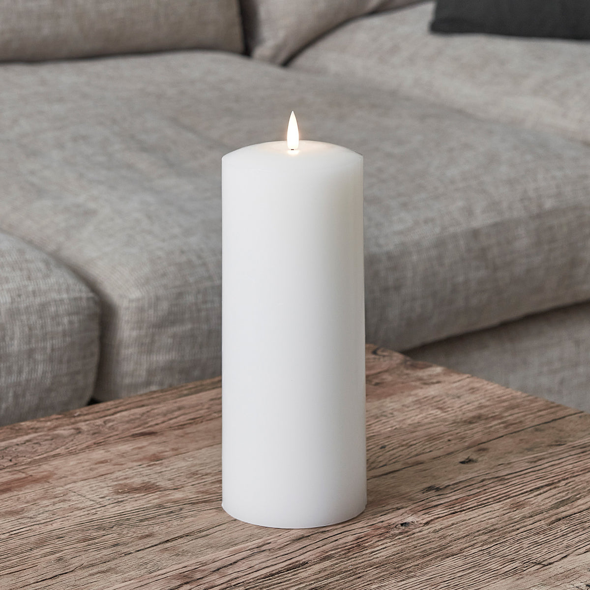 Weiße LED Stumpenkerze mit flackernder Flamme auf Holztisch vor grauem Sofa