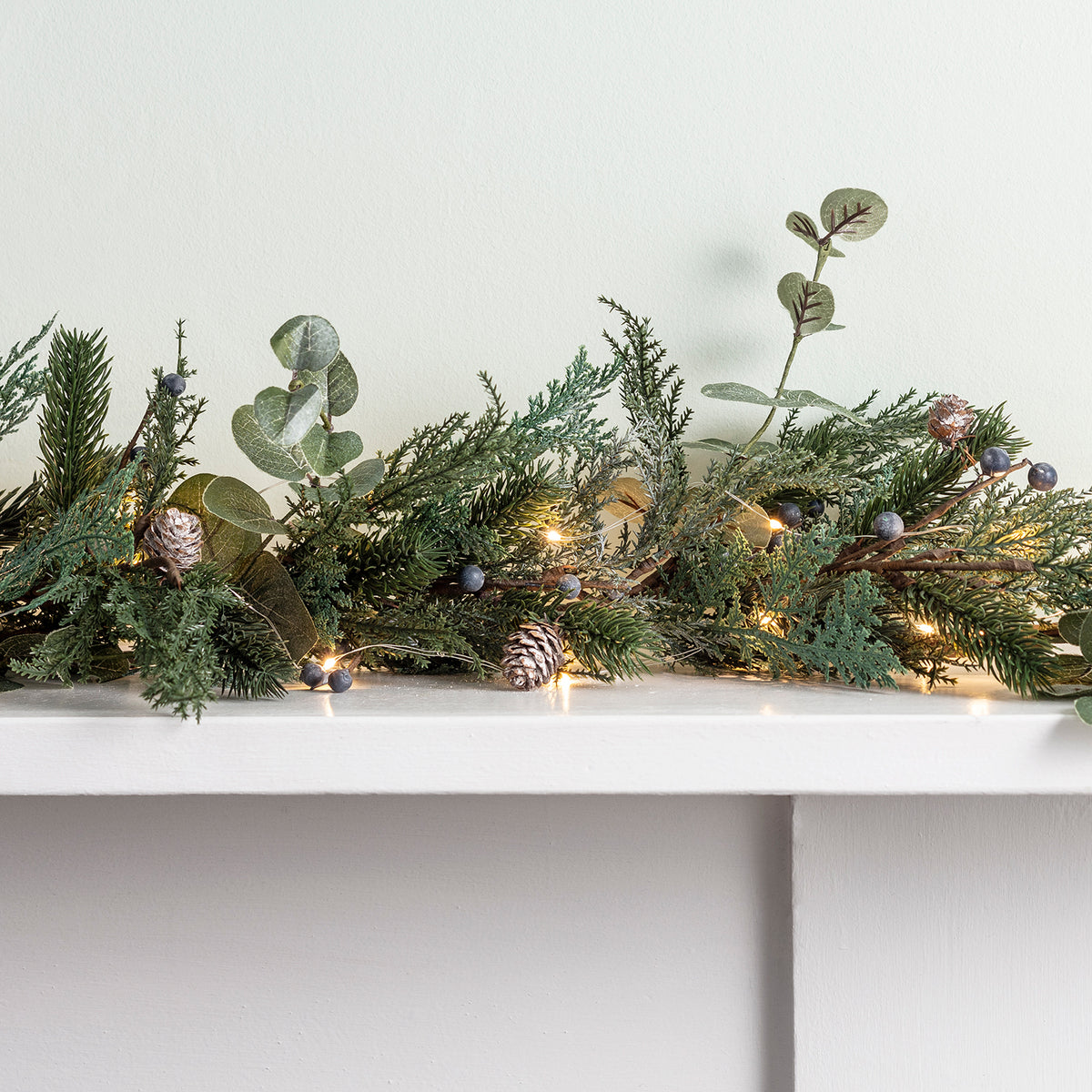 Nahaufnahme der Weihnachtsgirlande mit Tannenzweigen, Beeren, Eukalyptus und warmweißen Lichtern auf weißem Sims