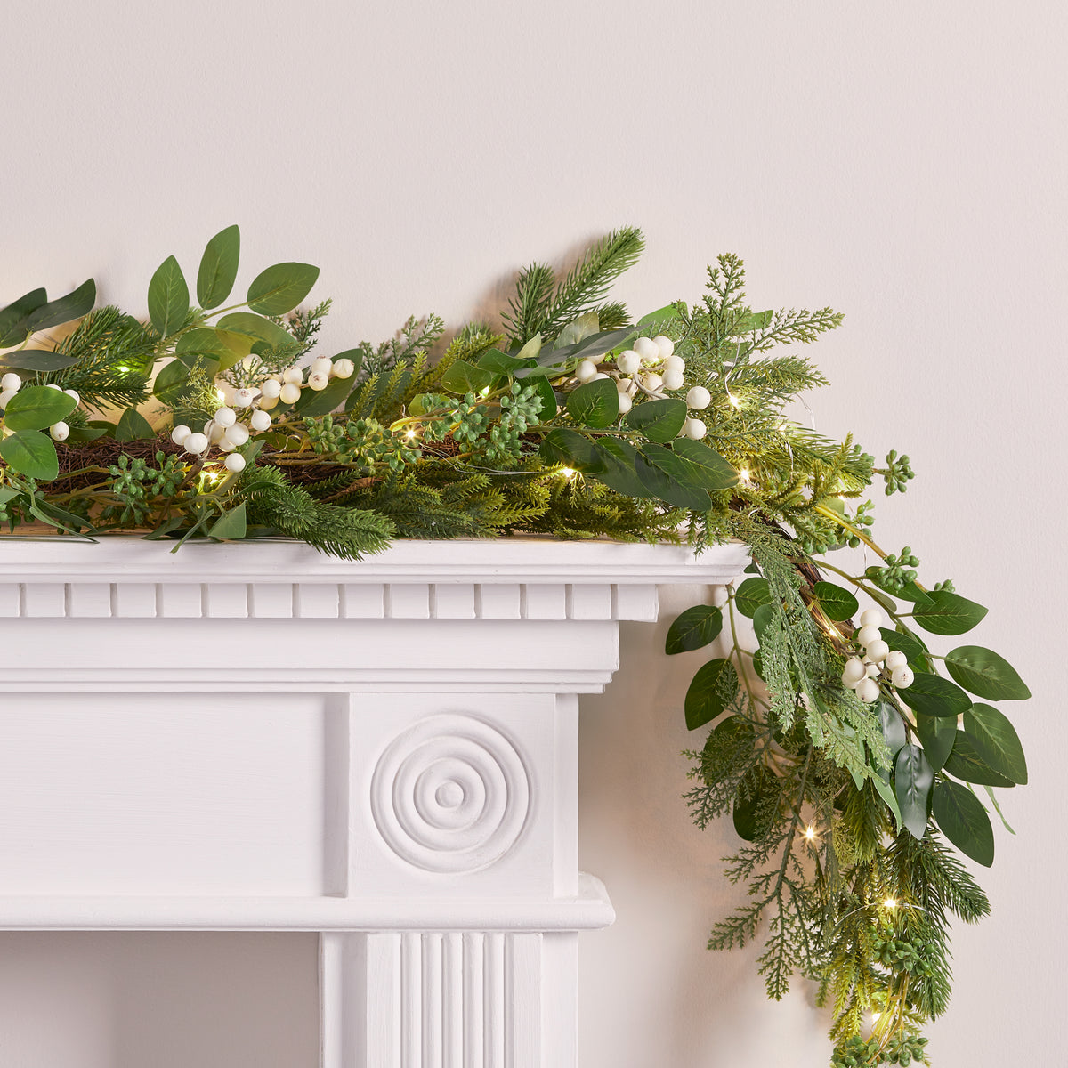 Nahaufnahme der grünen Weihnachtsgirlande mit weißen Beeren und warmweißen LED-Lichtern auf weißem Kaminsims