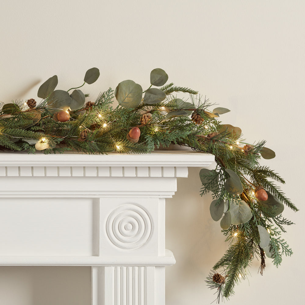 Weihnachtliche Girlande mit Eicheln, Tannenzapfen, grünen Blättern und Micro-Lichterkette auf weißem Kaminsims