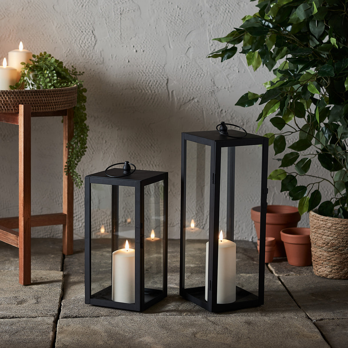 Zwei schwarze Laternen mit TruGlow Outdoor Kerzen auf Steinboden neben Holztisch und Pflanzen